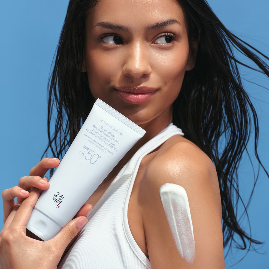 Kvindelig model med solcreme SPF 50 på skulderen, klar til udendørs aktivitet, mod en blå baggrund.